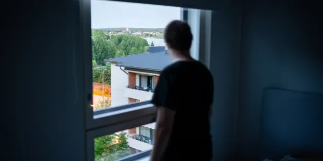 Ihminen katselee hämärässä huoneessa ikkunasta ulos lähiömaisemaa.